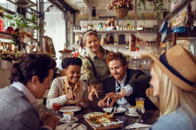 Un groupe de quatre amis souriants attablés dans un restaurant local chaleureux, servis par une serveuse bienveillante dans un cadre décoré de plantes et de bocaux colorés.
