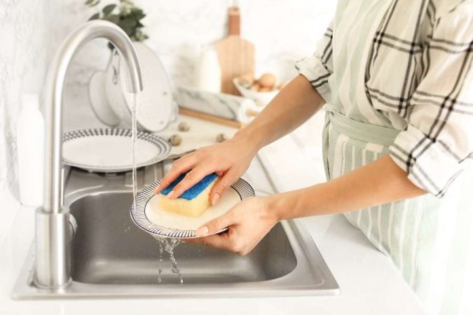 Une femme lave une assiette à l'aide d'une éponge sous le robinet ouvert d'un évier de cuisine en acier inoxydable.